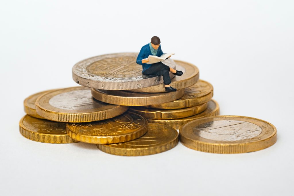 Eine winzige männliche Figur, die eine Zeitung liest und auf einem Haufen Münzen sitzt.